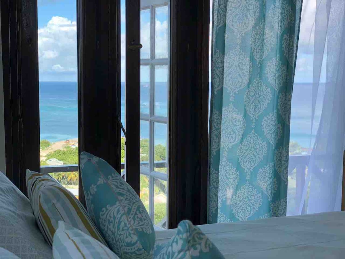 The image reveals a serene sleeping area, featuring a bed adorned with decorative pillows. Large windows framed by light blue curtains showcase panoramic views of the Caribbean Sea and the Atlantic Ocean, with clear skies and distant shoreline visible.