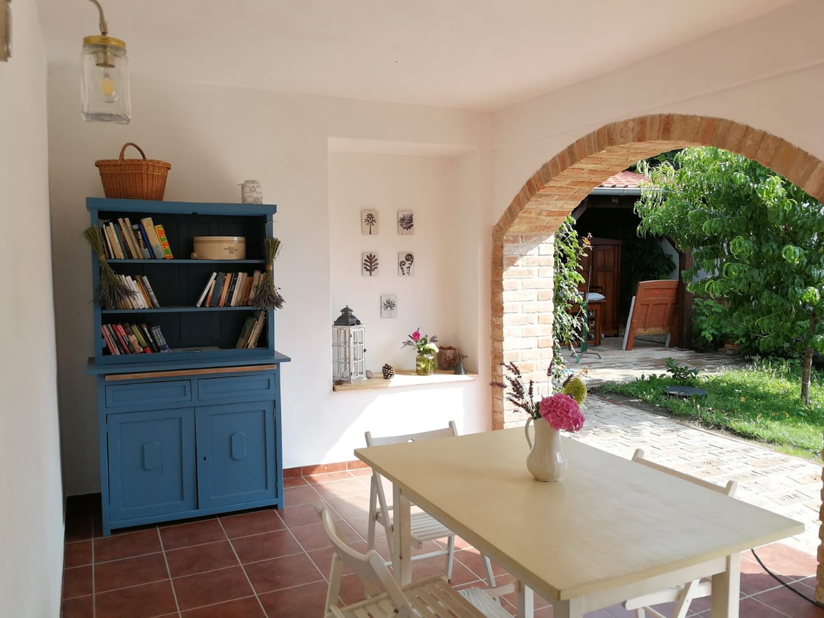 A cozy outdoor dining area features a light wooden table surrounded by white chairs. A blue bookcase filled with books stands nearby, accompanied by a decorative lantern and a small flower arrangement. Natural light filters in, highlighting a lush garden visible through an archway.