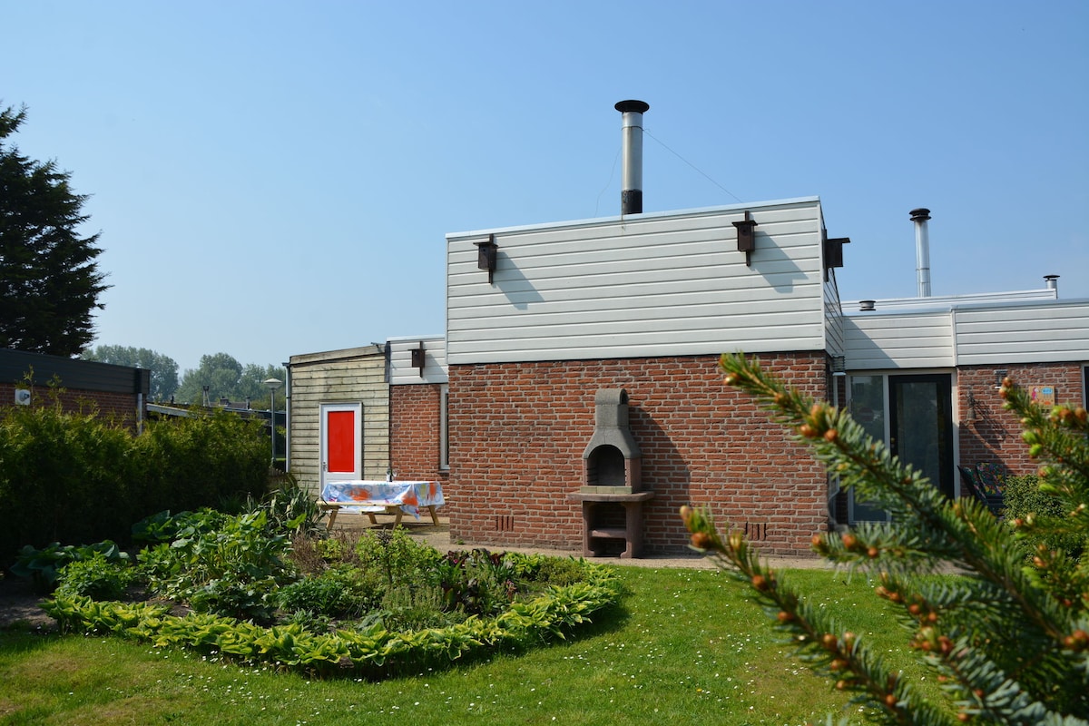 The exterior of the holiday home features a brick facade accented by modern cladding. A well-maintained garden surrounds the home, showcasing a circular flower bed. A barbecue grill is positioned near the garden, and a vibrant red door adds a pop of color to the view.