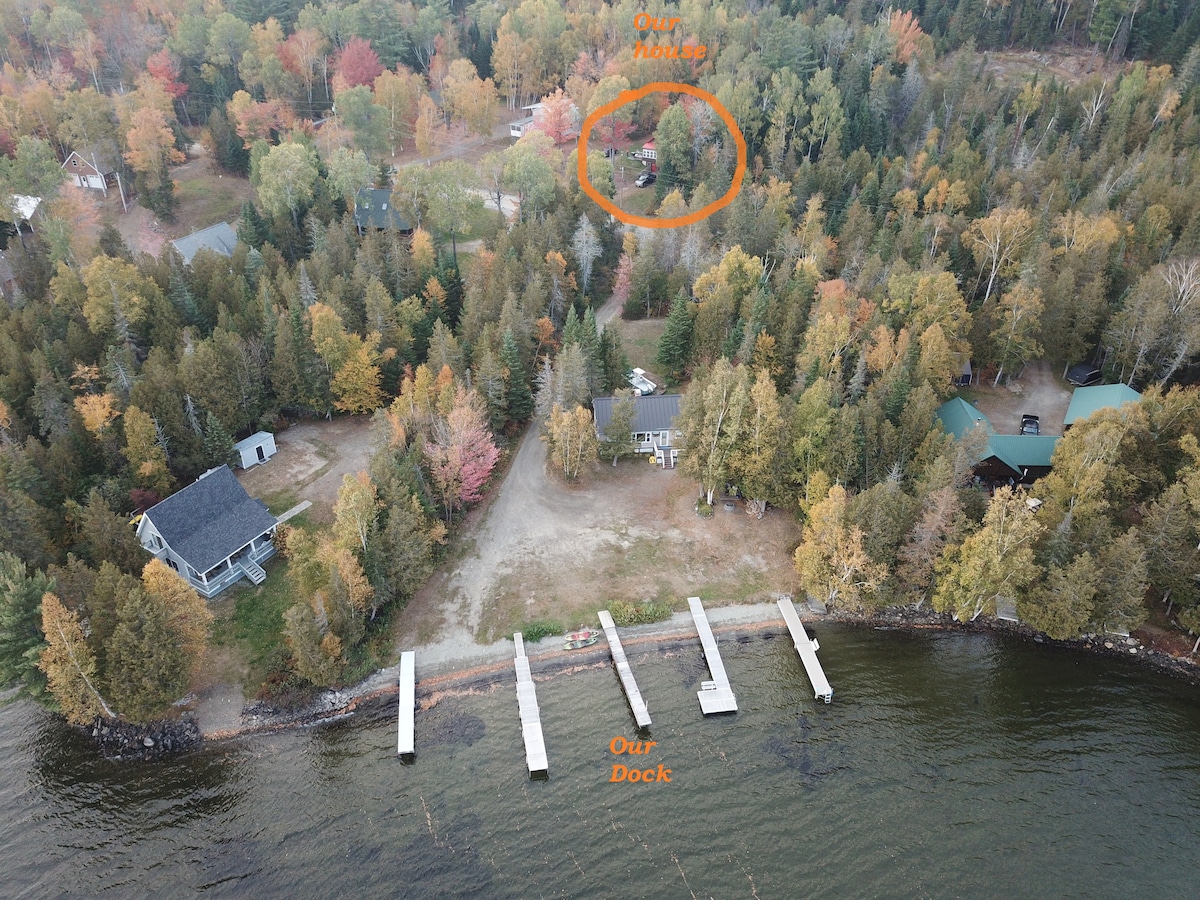 An aerial view showcases the property set amidst a lush landscape, with trees displaying autumn colors. The nearby dock provides multiple docking spaces along the calm water, while the house is positioned slightly elevated, offering views of the surroundings.