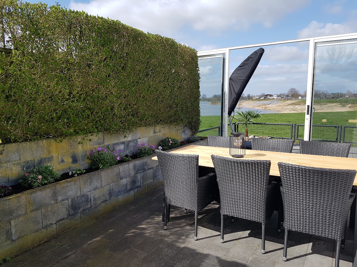 An outdoor dining area features a large wooden table surrounded by black wicker chairs. A tall, dark umbrella stands nearby for shade. Lush greenery serves as a natural privacy barrier, and flower beds line the wall, adding color to the serene lakeside view.