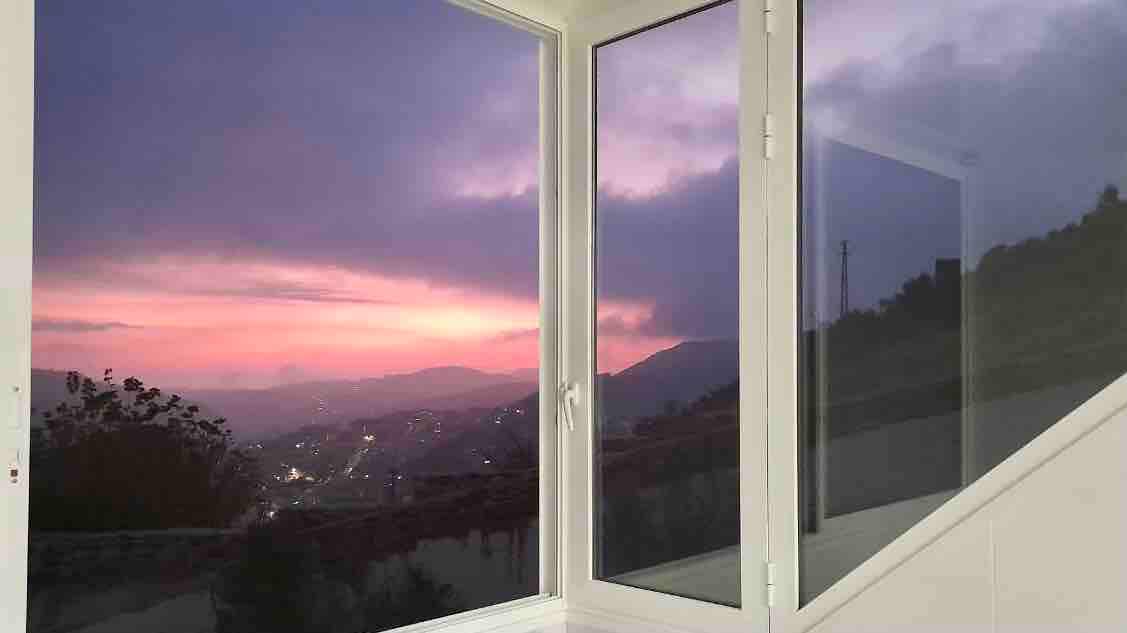 A spacious window offers a view of a serene landscape at dusk, showcasing a soft pink and purple sky. The surrounding hills are visible, with distant lights glimmering in the valley below.
