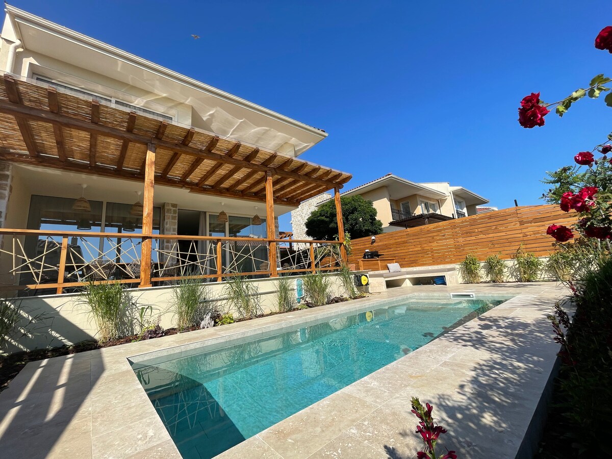 A private swimming pool is framed by a stone patio, surrounded by lush greenery and colorful flowers. A spacious covered terrace with wooden beams overlooks the pool and garden, providing a relaxing outdoor space. Clear blue skies serve as a backdrop to the villa.