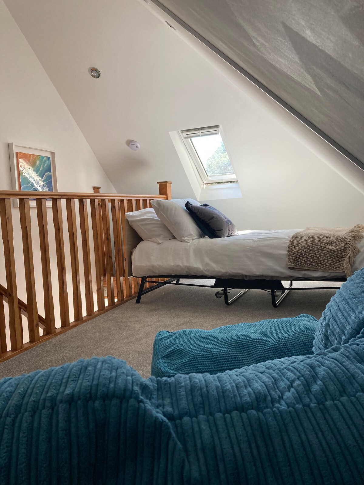 An inviting loft space is highlighted by a spacious bed with decorative pillows. A cozy beanbag chair in a soft blue is positioned in the foreground. Natural light filters through a small window, illuminating the sloped ceiling and wooden railing.