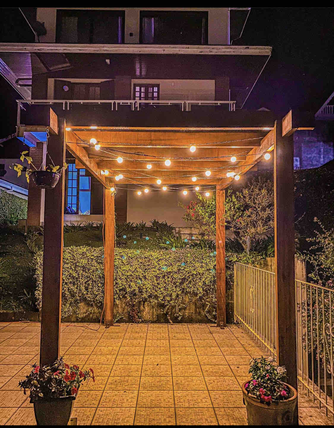 A wooden pergola is adorned with string lights, creating a warm ambiance. Flower pots are placed on the ground, alongside a paved patio area. Lush greenery is visible in the background, enhancing the tranquility of the outdoor space.