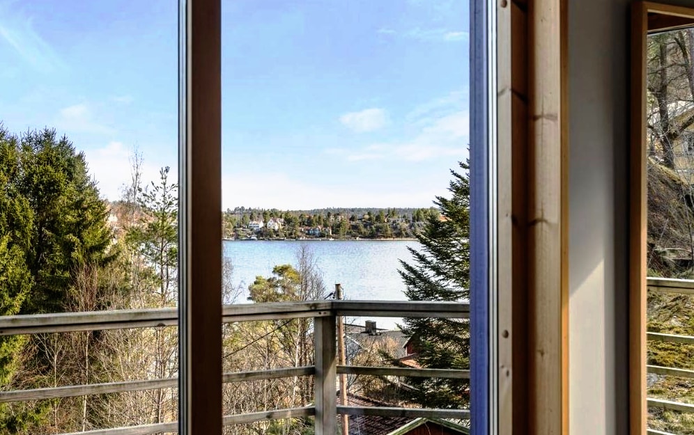 A view of the serene waterfront is framed by large glass windows, allowing natural light to illuminate the space. The scene showcases trees lining the shore, with a calm body of water in the distance, enhancing the connection to nature.