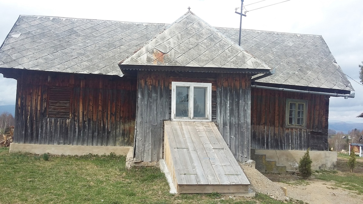 A rustic mountain house is featured with a wooden exterior, showcasing natural wood tones. A gently sloped ramp leads to the entrance, facilitating accessibility. Windows framed in green add a charming touch, and the surrounding grassy area is complemented by distant mountainous views.