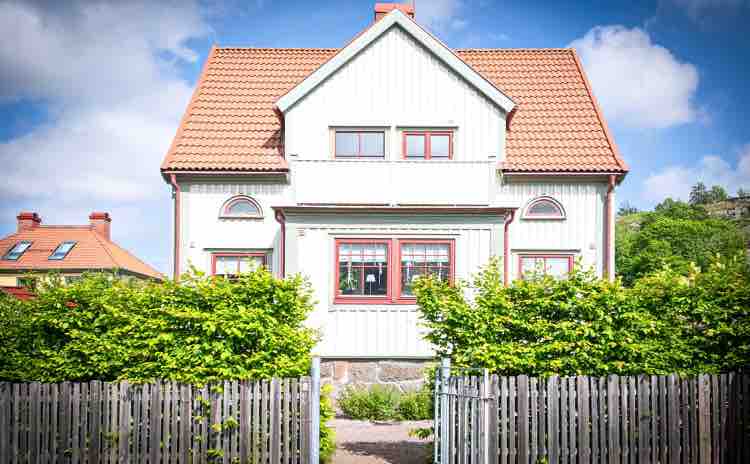 The exterior of a two-story villa is shown, featuring a white façade with red accents and a tiled roof. The house is framed by green bushes and a wooden fence, creating an inviting approach. Large windows illuminate the front, reflecting a clear blue sky.