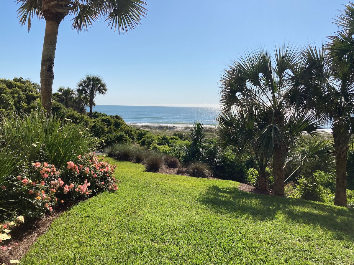 A view of lush green landscaping featuring palm trees and flowering bushes. The vibrant colors contrast with the sandy beachfront and the Atlantic Ocean beyond, showcasing a serene and natural coastal setting under a clear blue sky.