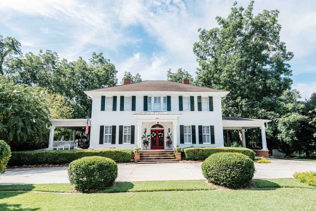 The historic home features a classic white exterior with green shutters, complemented by a welcoming red front door. Surrounding the entrance are lush green shrubs and a well-maintained yard. A covered porch extends along the front, providing shaded seating options.