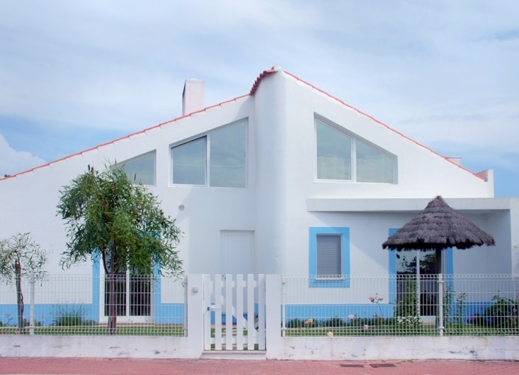 A modern white house features an angular roof design and multiple large windows, allowing ample natural light. The exterior is complemented by a blue color accent, while a shaded outdoor area is provided by a thatched roof. A fenced garden area is visible.