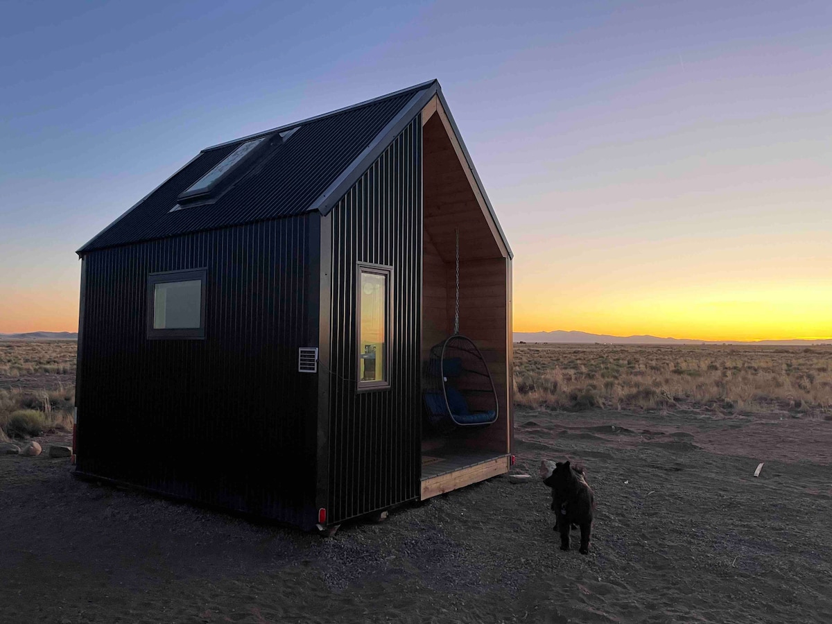 The tiny house features a sleek black exterior with large windows that reflect the sunset. A hanging chair is situated on the porch, providing a cozy spot to enjoy the view. An open landscape extends around the home, highlighted by natural grasses.