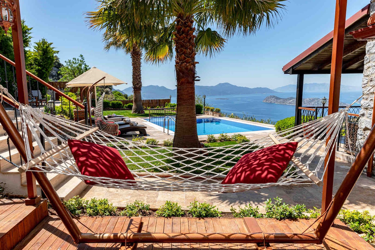 A spacious hammock, adorned with red cushions, is positioned between sturdy posts on a wooden decking. In the background, a clear pool reflects the sun, surrounded by lush greenery and palm trees, with serene sea views extending to distant islands.