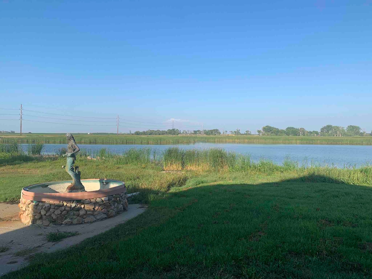 A serene outdoor landscape features a stone fountain with a statue, set against a backdrop of calm water. Lush green grass surrounds the fountain, while tall grasses frame the peaceful water's edge. The horizon showcases a clear blue sky and distant trees.