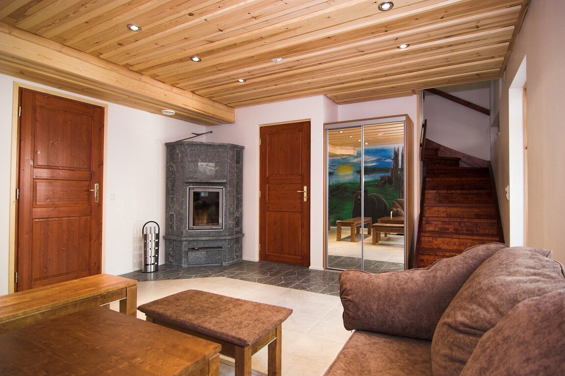 A spacious living area features a wooden ceiling and a cozy sofa. A stone fireplace adds warmth, while glass doors lead to a welcoming space with scenic artwork. A wooden staircase provides access to the upper level, enhancing the home's inviting layout.