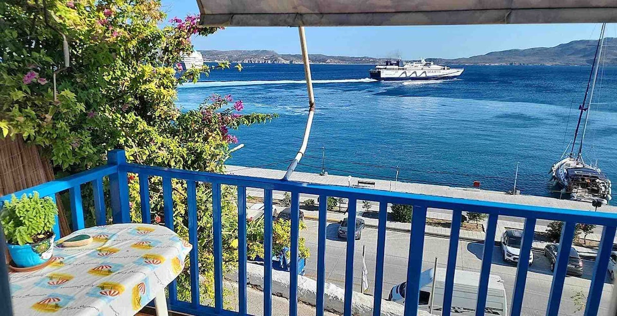 A balcony reveals bright blue railings with a small, circular table adorned with a cheerful pattern. The sea glimmers under sunlight, displaying various shades of blue, while a large ship passes by. Green foliage frames the scene, enhancing the coastal view.