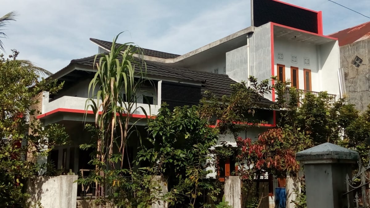The exterior of the house is shown, featuring a two-story structure with a modern design. The facade is adorned with a mix of light and dark colors, complemented by greenery from surrounding plants. Large windows invite natural light, highlighting the welcoming atmosphere.
