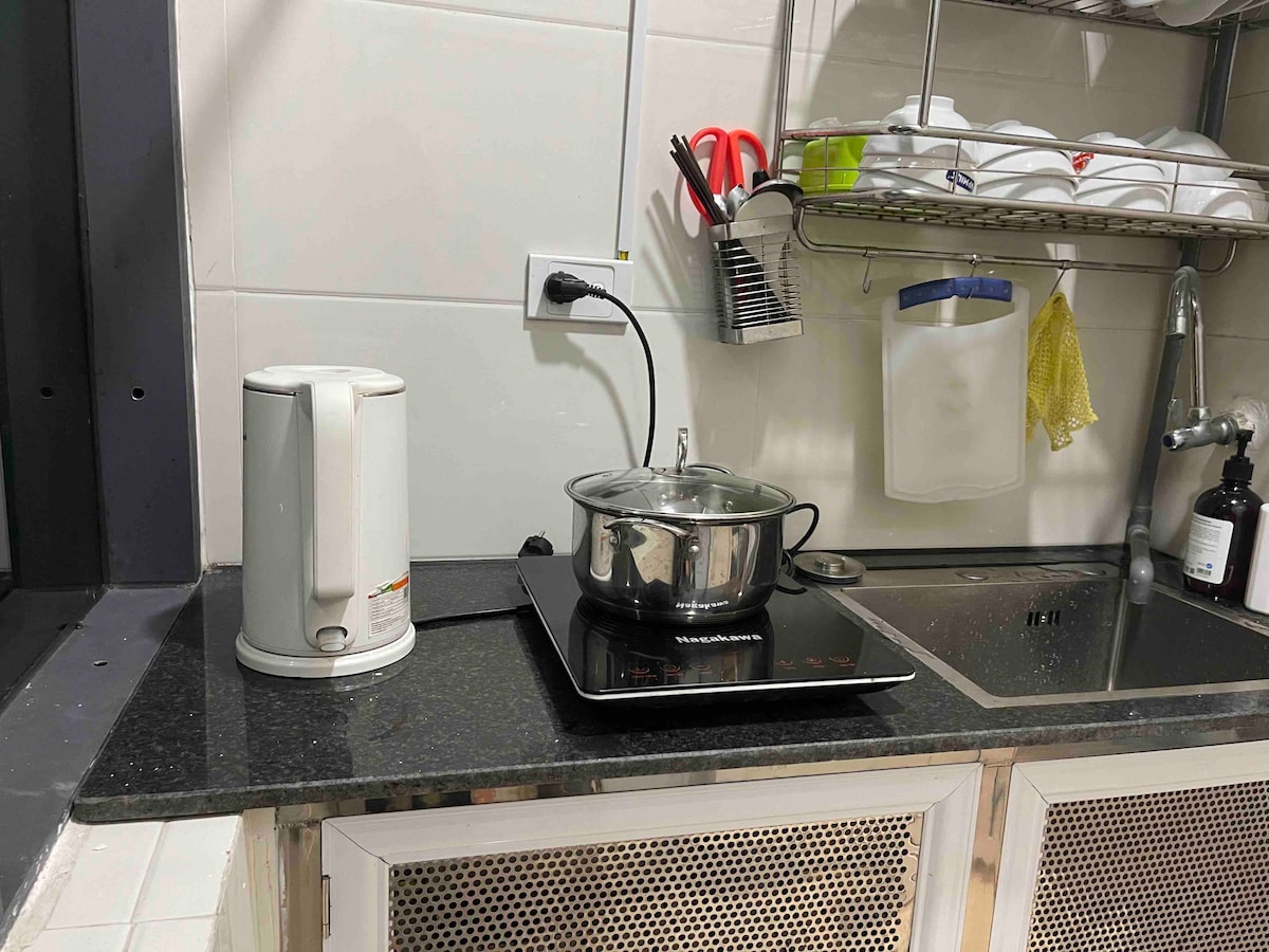 The kitchen area includes a countertop with an electric kettle and a saucepan on an induction stove. A wire rack above holds various utensils. A sink is visible with dish soap nearby, providing essential conveniences for meal preparation.