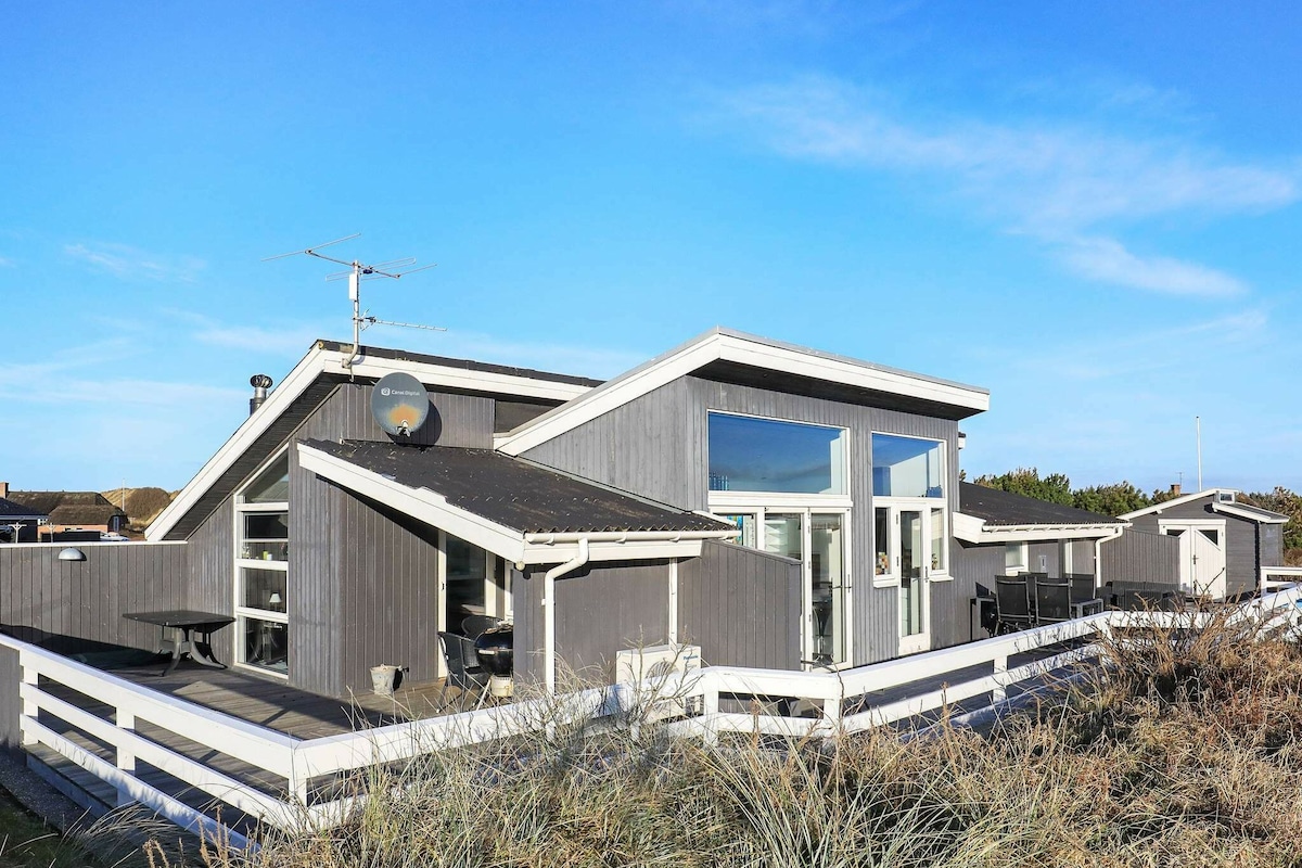 A modern holiday home is depicted, featuring a unique architecture with multiple slanted roofs. Large windows provide ample natural light, and outdoor seating is visible. The house is surrounded by a well-maintained fence, with sandy dunes and greenery in the foreground under a clear blue sky.