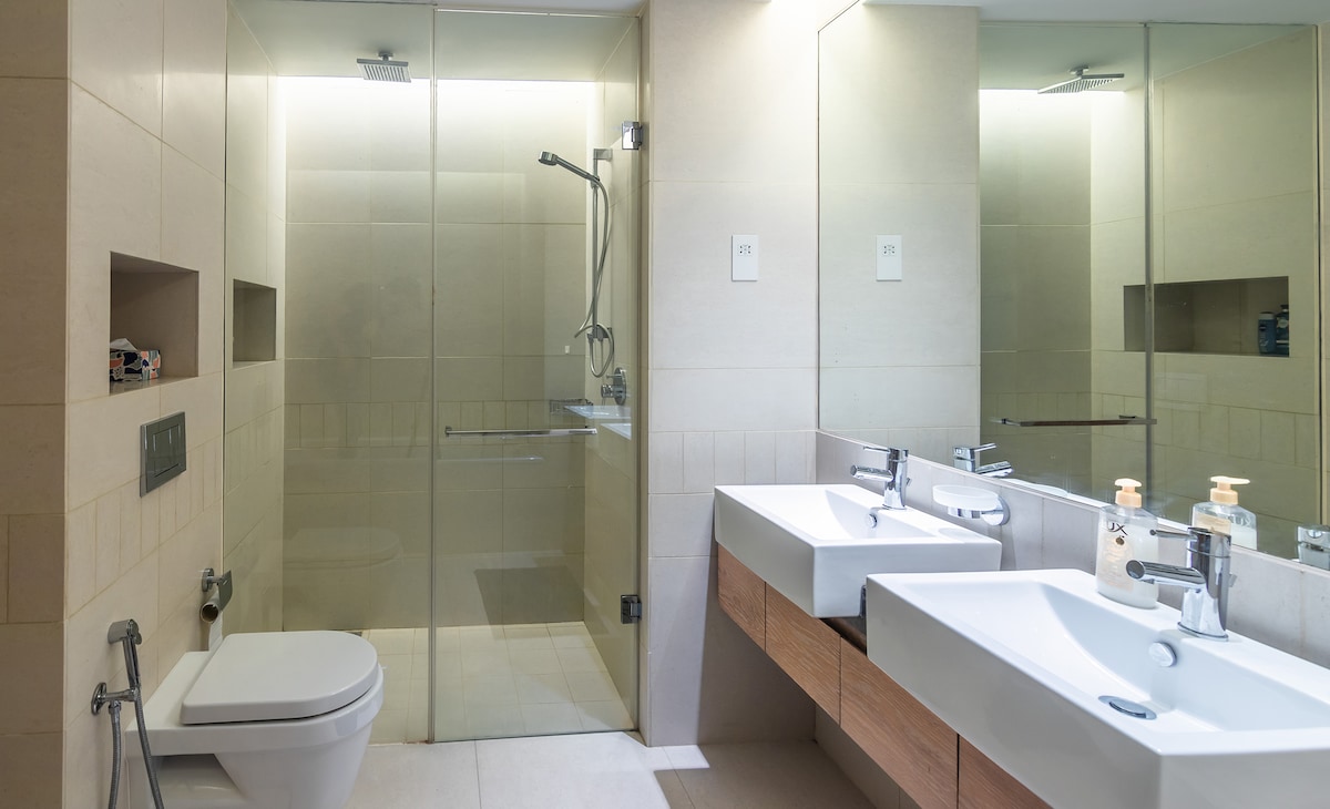 A modern bathroom is displayed, featuring a glass-enclosed shower and two sleek sinks with wooden cabinetry. Natural light reflects off the mirrors, enhancing the spacious feel. A toilet is positioned discreetly, and storage areas are integrated into the design for convenience.