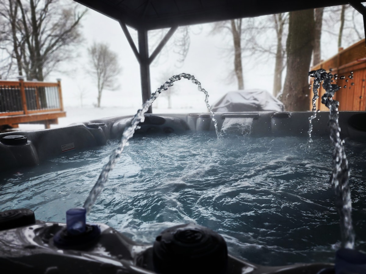 A hot tub is situated beneath a covered patio, featuring jets that create gentle water splashes. The serene winter landscape is visible in the background, with snow lightly covering the ground and trees. The atmosphere suggests a private and relaxing retreat.