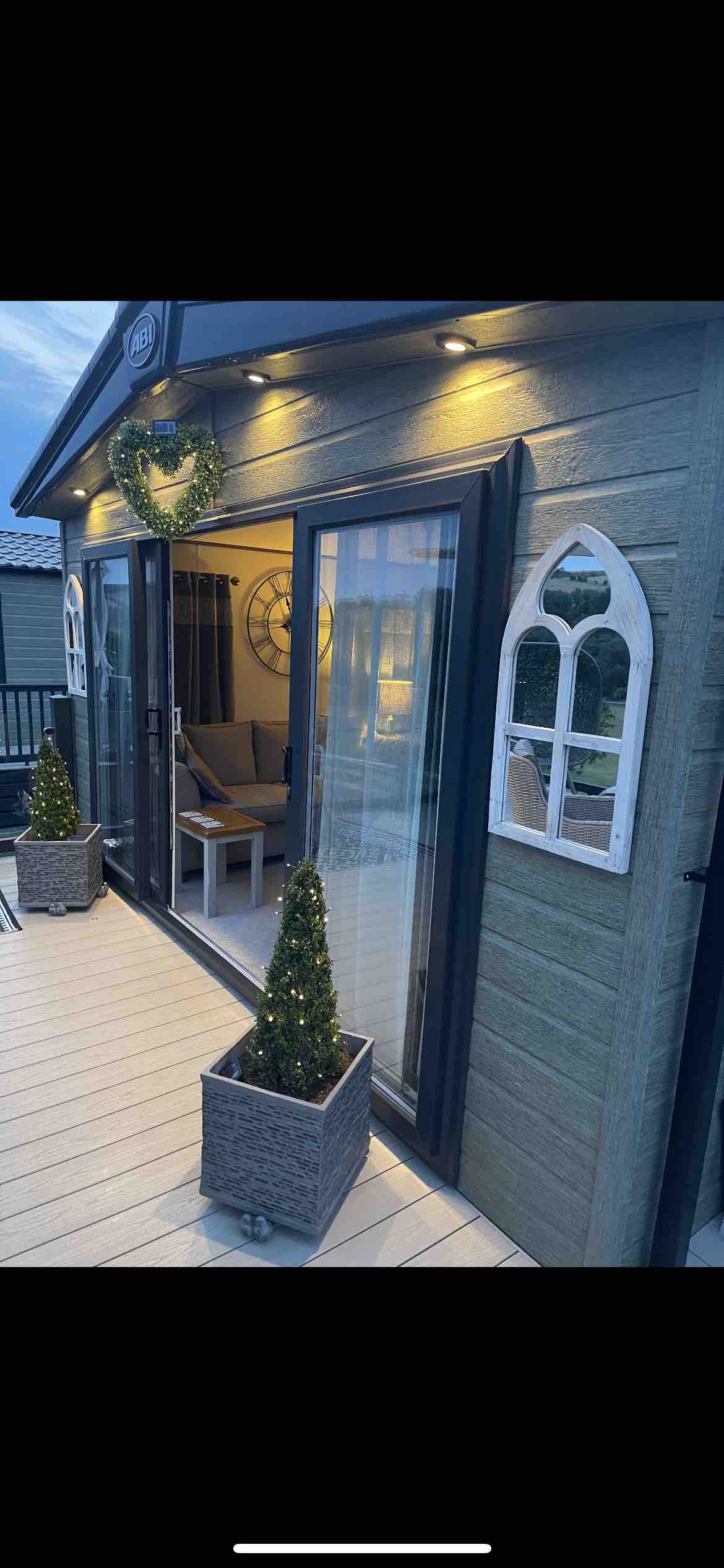 The exterior of the mini luxury lodge features large glass doors that invite natural light. Potted evergreen plants are placed alongside the deck, enhancing the welcoming entry. Soft lighting illuminates the porch, creating a cozy ambiance for relaxation.