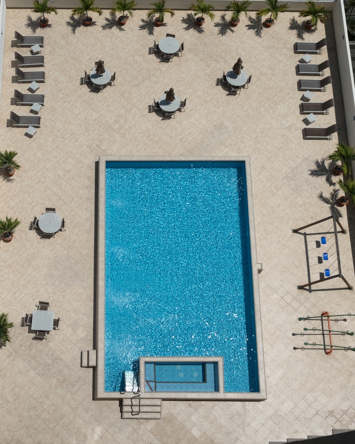 An aerial view of a swimming pool is presented, surrounded by spacious tiled areas. Multiple lounge chairs and tables are arranged around the pool, with palm trees providing hints of greenery. The pool features a clear blue water surface, and there is a spa area integrated on one side.