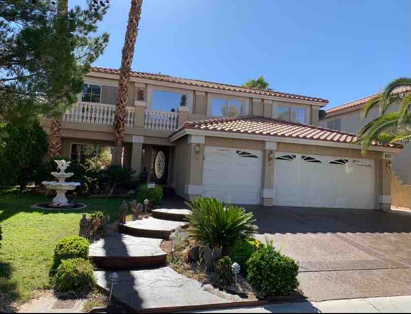 A spacious two-story house with a tile roof is set in a well-maintained yard featuring palm trees and various shrubs. A decorative fountain is visible in the front garden, and the driveway leads to a two-car garage with arched doors.