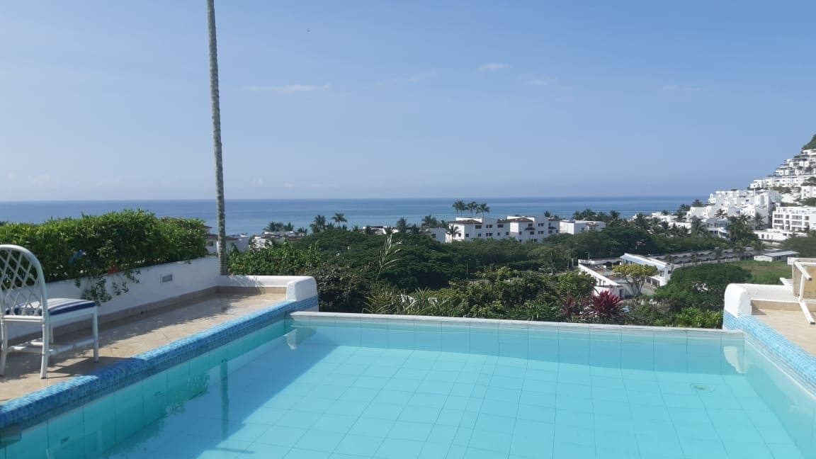 A serene view overlooking a private infinity pool captures vibrant greenery and coastal waters. The clear blue sky complements the tranquil atmosphere, while comfortable loungers and a single chair are positioned near the pool edge, inviting relaxation.