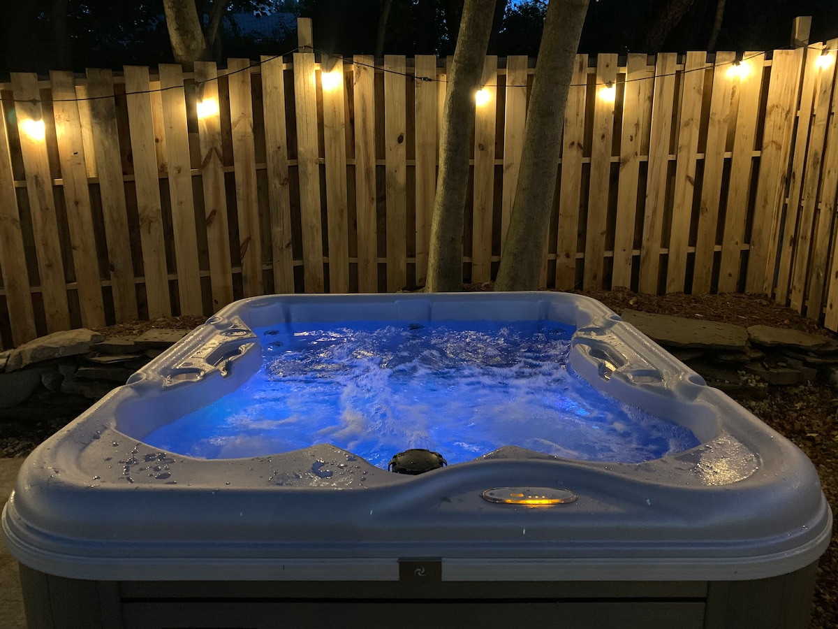 A bubbling hot tub sits illuminated by soft blue lighting, surrounded by a wooden fence. Softly lit string lights hang above, creating a serene outdoor atmosphere for relaxation.