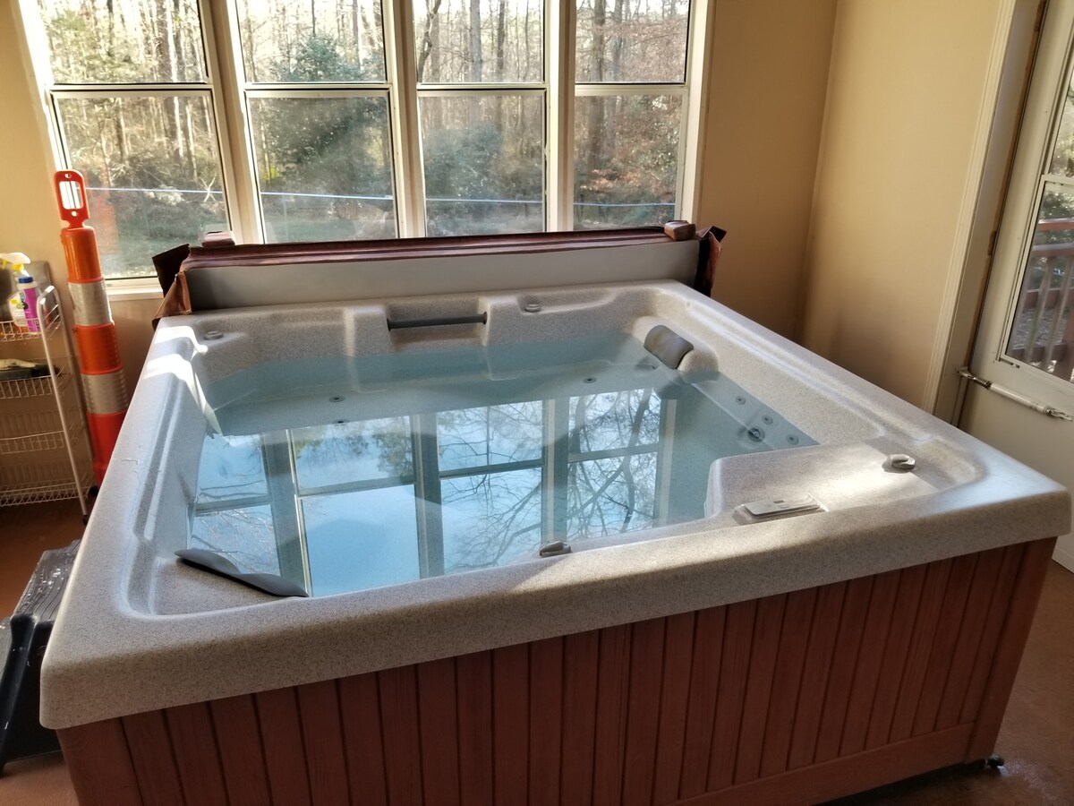 A hot tub is positioned in a sunlit room with large windows, allowing natural light to reflect off the water. The tub features a smooth surface and wooden paneling, surrounded by a serene landscape view outside the windows.