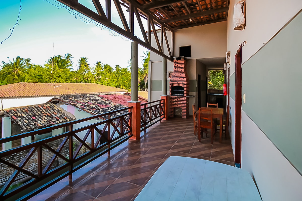 A spacious balcony is highlighted by wooden railings, offering views of surrounding trees and rooftops. A small dining table, an outdoor grill, and a flat-screen TV are visible, creating a comfortable space for relaxation. Natural light enters from the open area, enhancing the inviting environment.