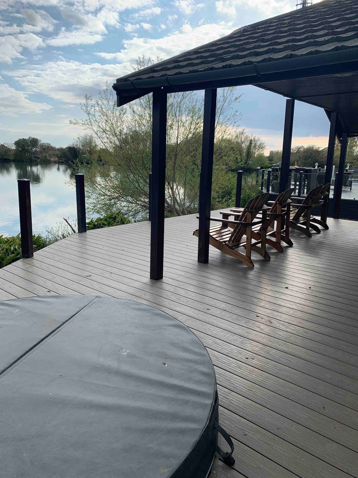 A private deck offers seating for relaxation, featuring several wooden chairs. A hot tub is present, partially covered, overlooking a serene lake surrounded by greenery. The calm water reflects the sky, adding to the tranquil setting.