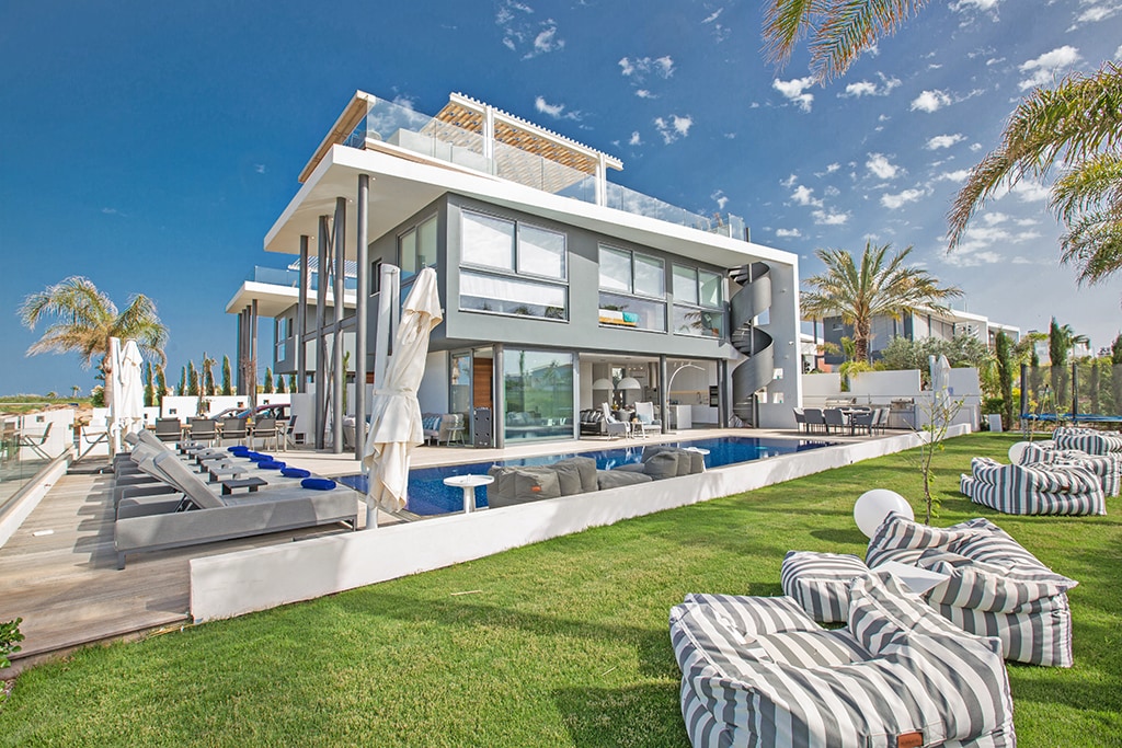 The modern villa features a spacious exterior with a large private pool, surrounded by a well-maintained lawn. Sun loungers line the poolside, and cozy beanbags are scattered throughout the grass area. Large windows showcase a bright, airy interior, with palm trees enhancing the tropical ambiance.