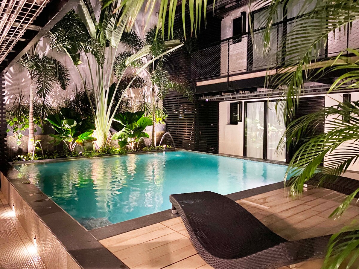 A serene pool area is surrounded by lush tropical plants, illuminated softly by night lighting. The tranquil water sparkles, reflecting the greenery. Lounge chairs are positioned near the water's edge, offering a relaxing space for guests.