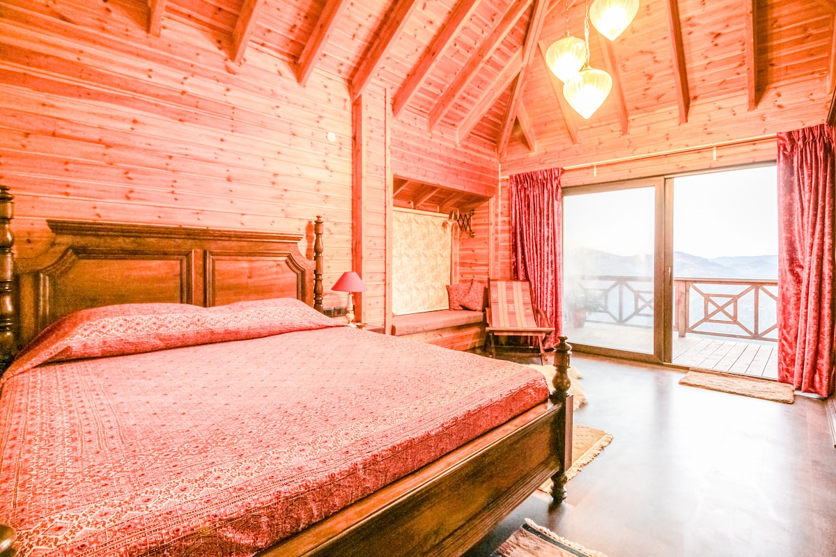 A wooden chalet room features a spacious bed adorned with a patterned maroon spread. Large glass doors open to a deck, providing views of the surrounding landscape. Pendant lights hang from the vaulted ceiling, and a cozy seating area is positioned to the side.