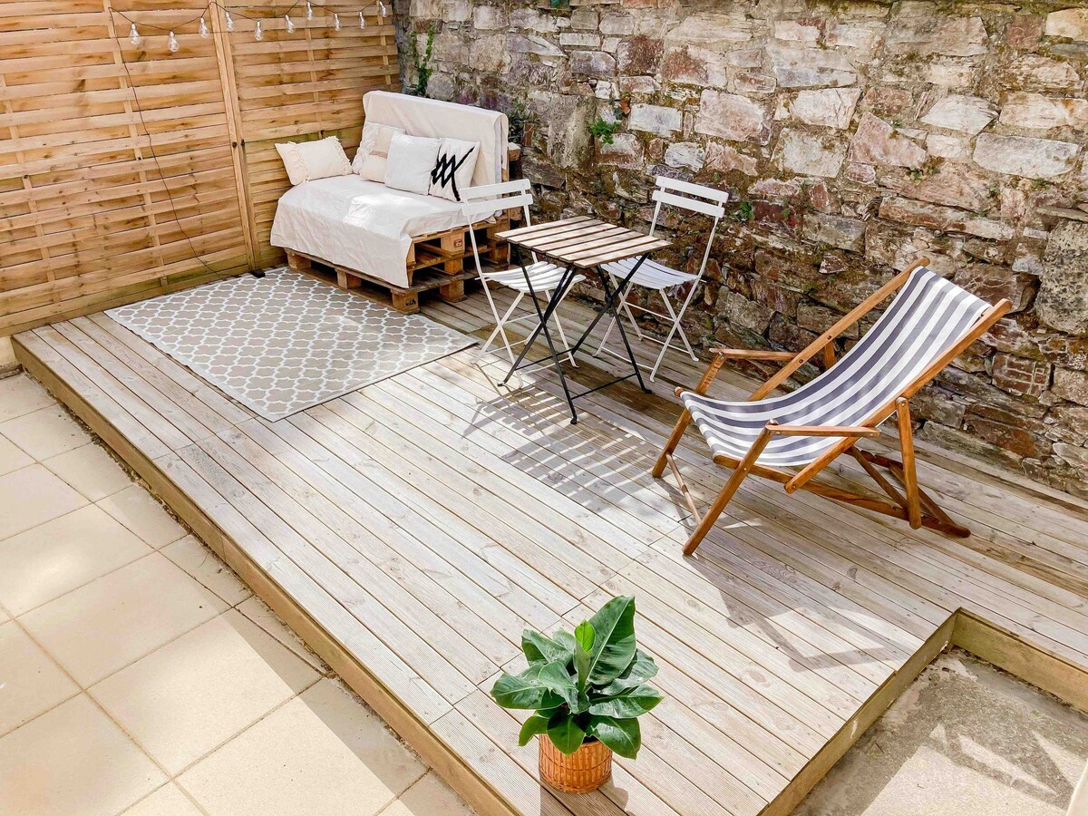 A wooden terrace features a small table with two chairs and a lounge chair, offering a cozy outdoor space. A light-colored couch made from pallets is positioned against a stone wall, complemented by a patterned rug and a potted plant for a touch of greenery.
