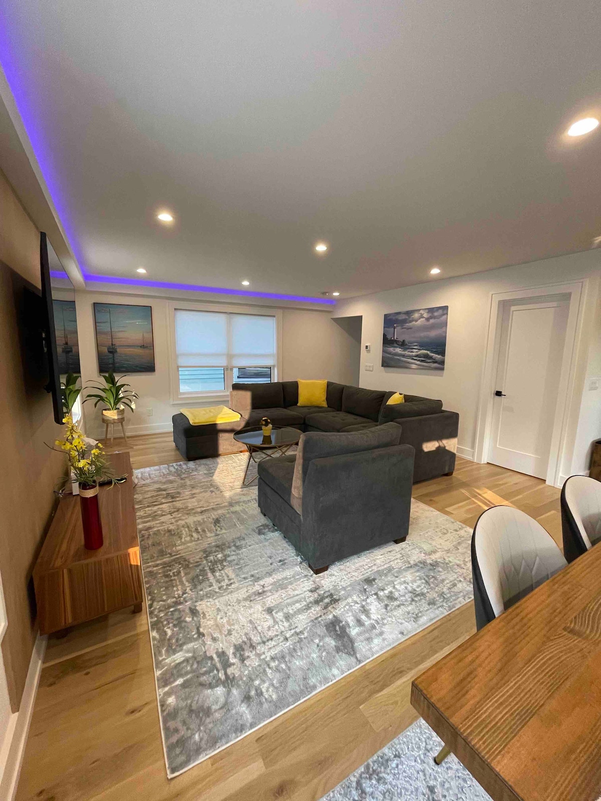 A spacious living room is depicted, featuring a large sectional sofa arranged around a decorative area rug. Two framed landscape images adorn the walls, and recessed lighting enhances the modern ambiance. Natural light enters through a partially covered window, contributing to a calming atmosphere.