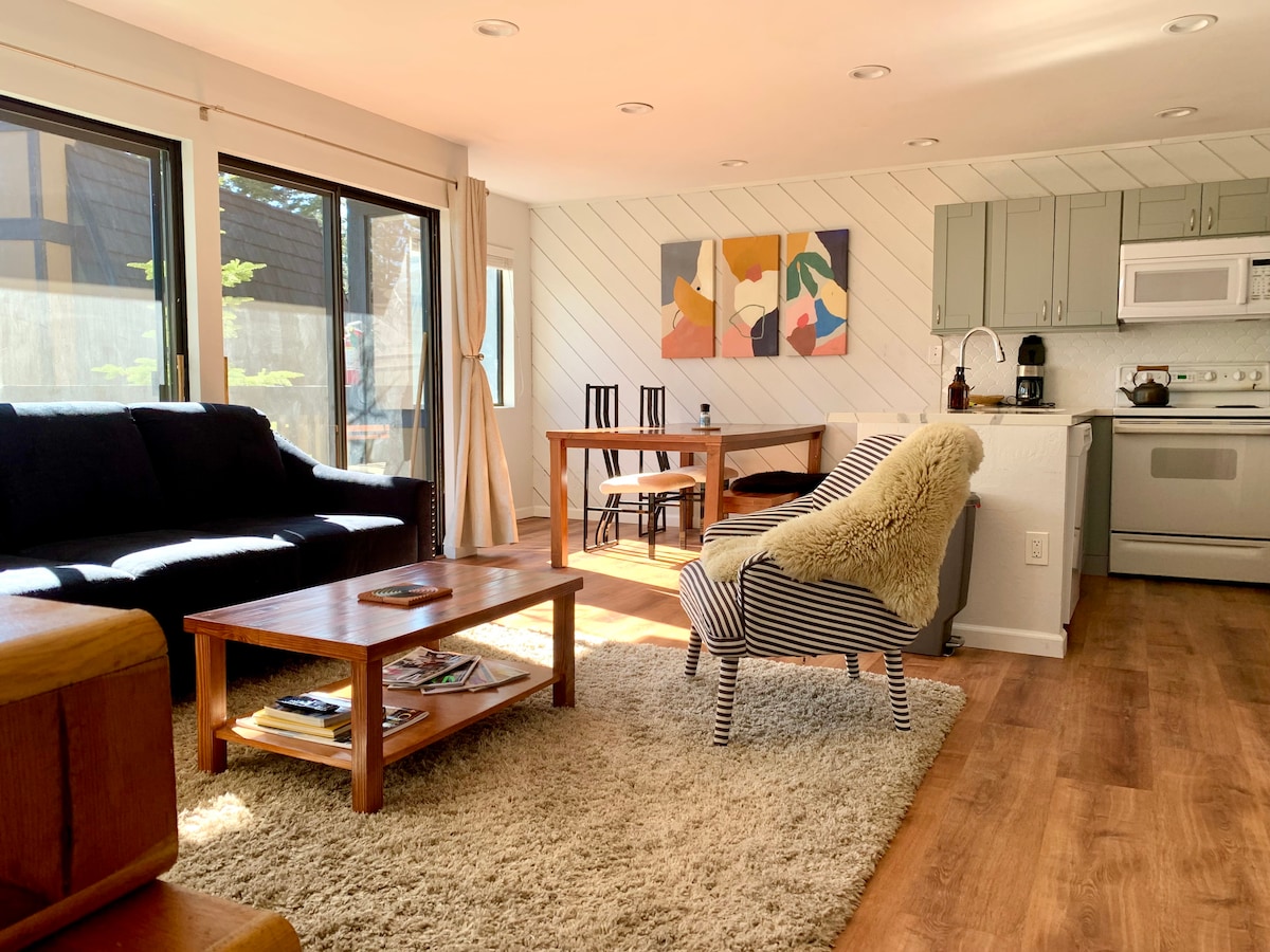 A bright living area features a dark sofa and a striped armchair, both positioned around a wooden coffee table. A dining table with chairs is visible nearby, while large windows allow natural light to fill the space. Colorful wall art adds a modern touch.