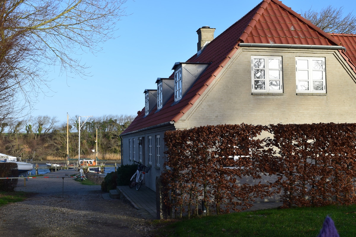 A charming two-story building is seen from the side, featuring a red-tiled roof and large windows. The surrounding area includes greenery and a glimpse of waterfront, suggesting a peaceful, natural setting ideal for relaxation.