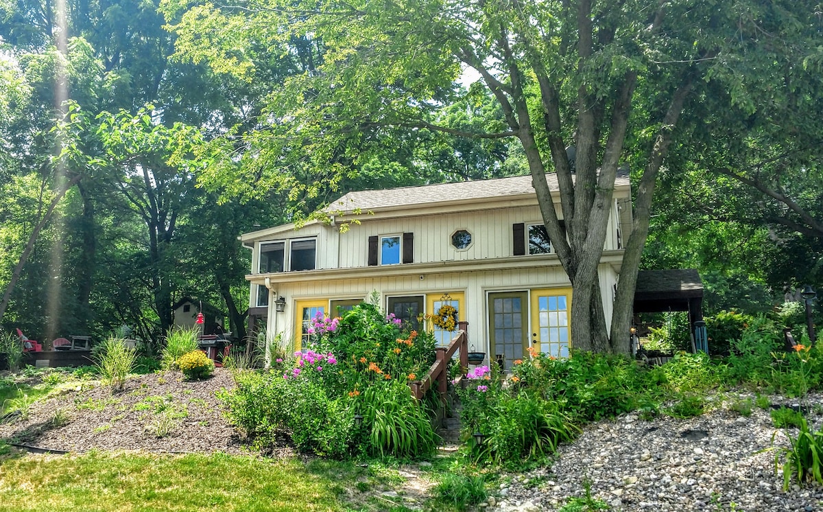 The cottage's exterior features a welcoming facade adorned with colorful flowers and greenery. A spacious deck is bordered by stone pathways, while large windows allow natural light to flood the living spaces. Mature trees provide shade in the surrounding area.