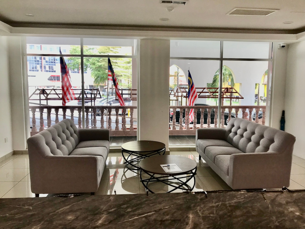 A bright lounge area is shown with two gray tufted sofas facing a round coffee table. Large windows allow natural light to enter, offering a view of outdoor seating adorned with flags. The modern design is complemented by polished tile flooring.