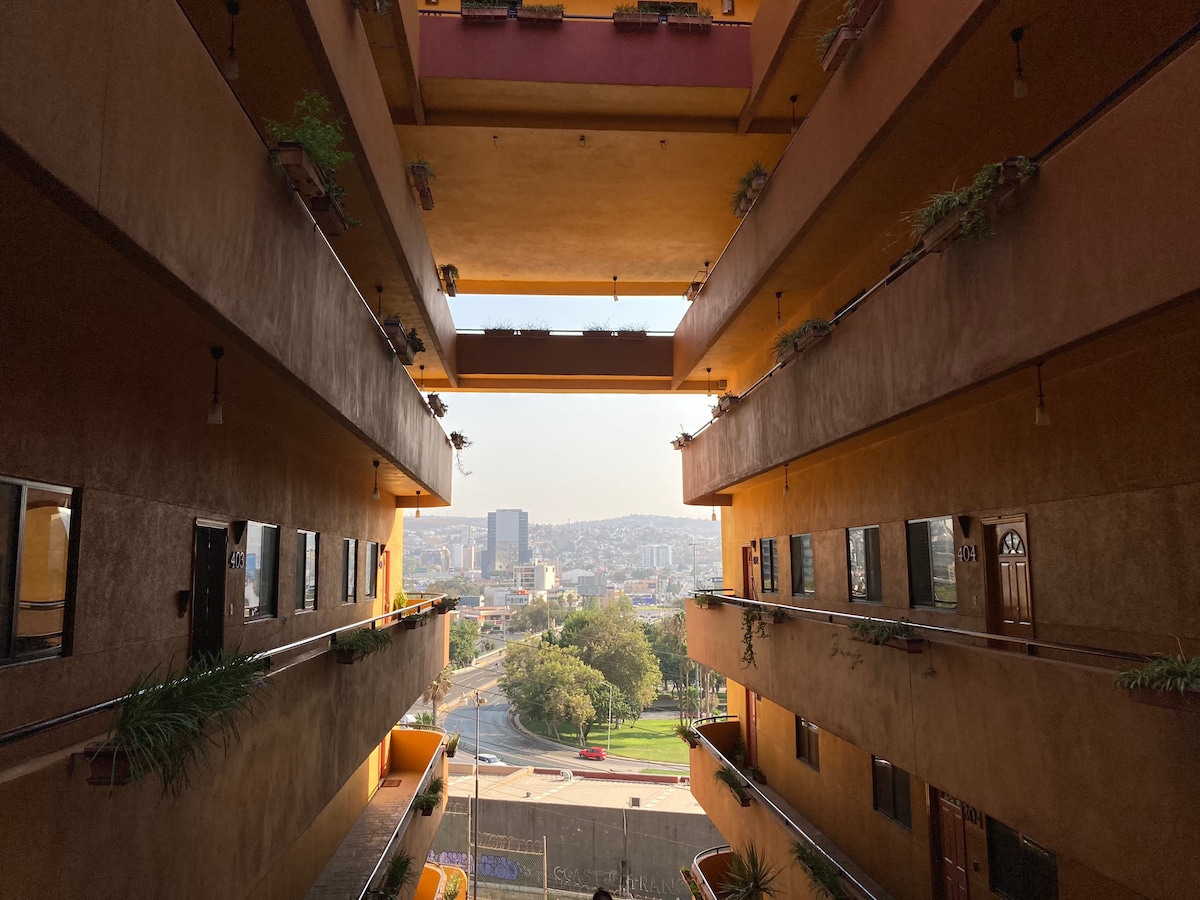 The view from the open corridor reveals a vibrant neighborhood with greenery and city buildings beyond. Natural light floods the space, highlighting the textured walls and hanging plants along the balconies. The structure features multiple levels, characterized by spacious walkways and doors leading to various apartments.