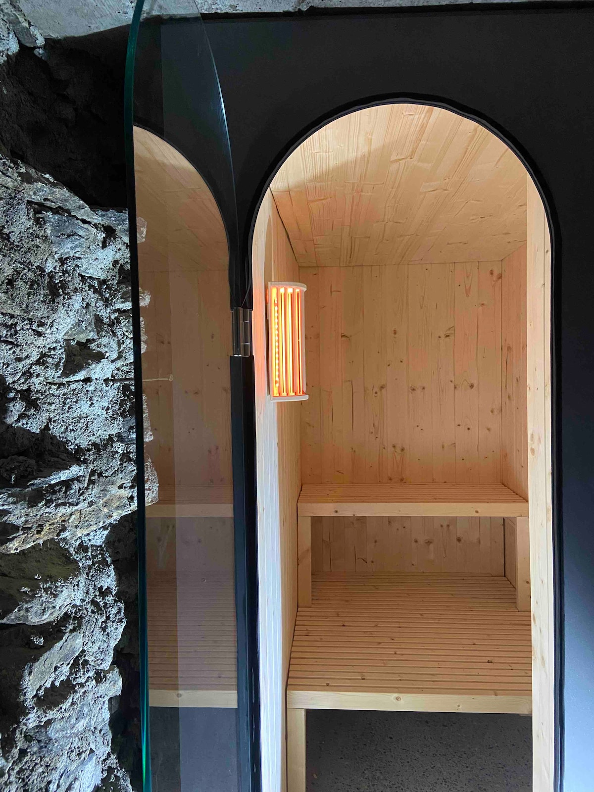 A modern sauna space features smooth wooden benches and paneling, creating a warm environment. Soft lighting casts a gentle glow, while the glass entrance adds an open feel. The textured stone wall contrasts with the sleek design, enhancing the serene atmosphere.