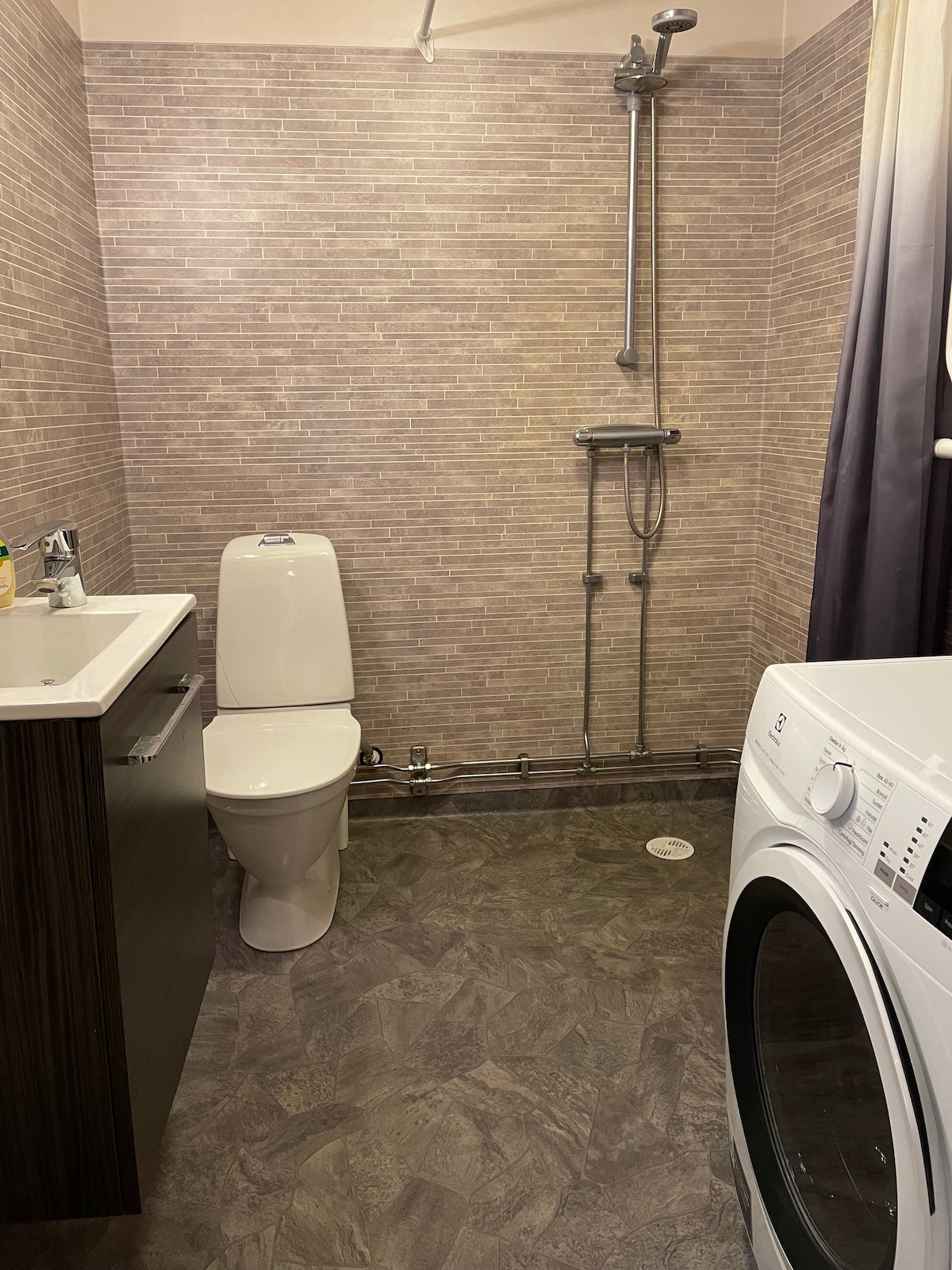 A modern bathroom features a white toilet positioned beside a sleek vanity with storage. A walk-in shower is highlighted by polished fixtures, and a washing machine occupies a corner. Textured beige tiles cover the walls, complementing the dark, patterned flooring.