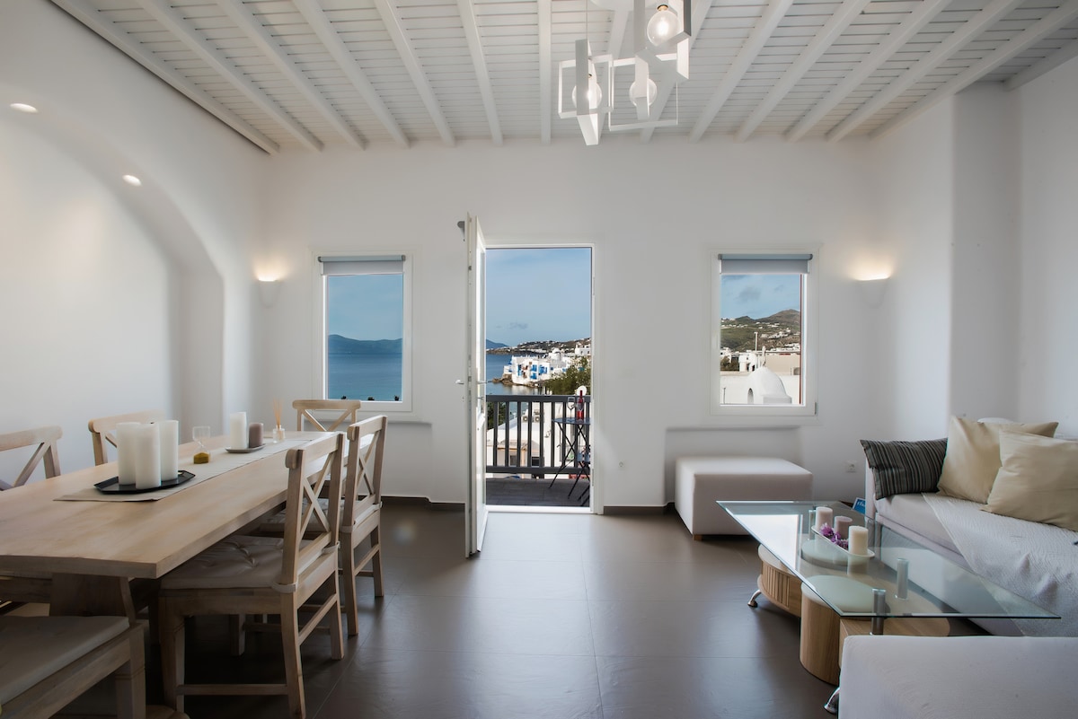 A bright living space features a wooden dining table with candles arranged on it. A glass coffee table sits in front of a comfortable sofa. Large windows allow natural light to fill the area, showcasing views of the sea and nearby buildings.