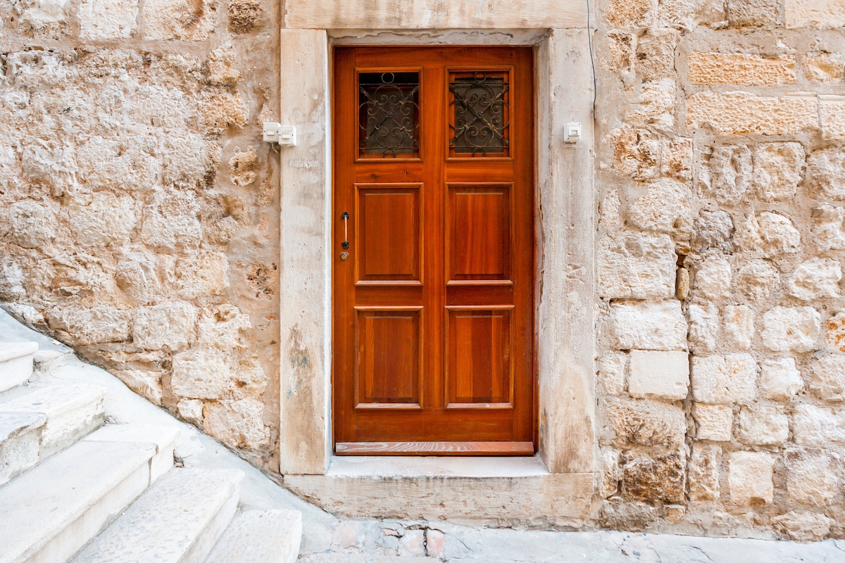A wooden door with a double paneled design is framed by a textured stone wall. The door features intricate glass detailing at the top and is bordered by light-colored stone steps that lead up from the street.