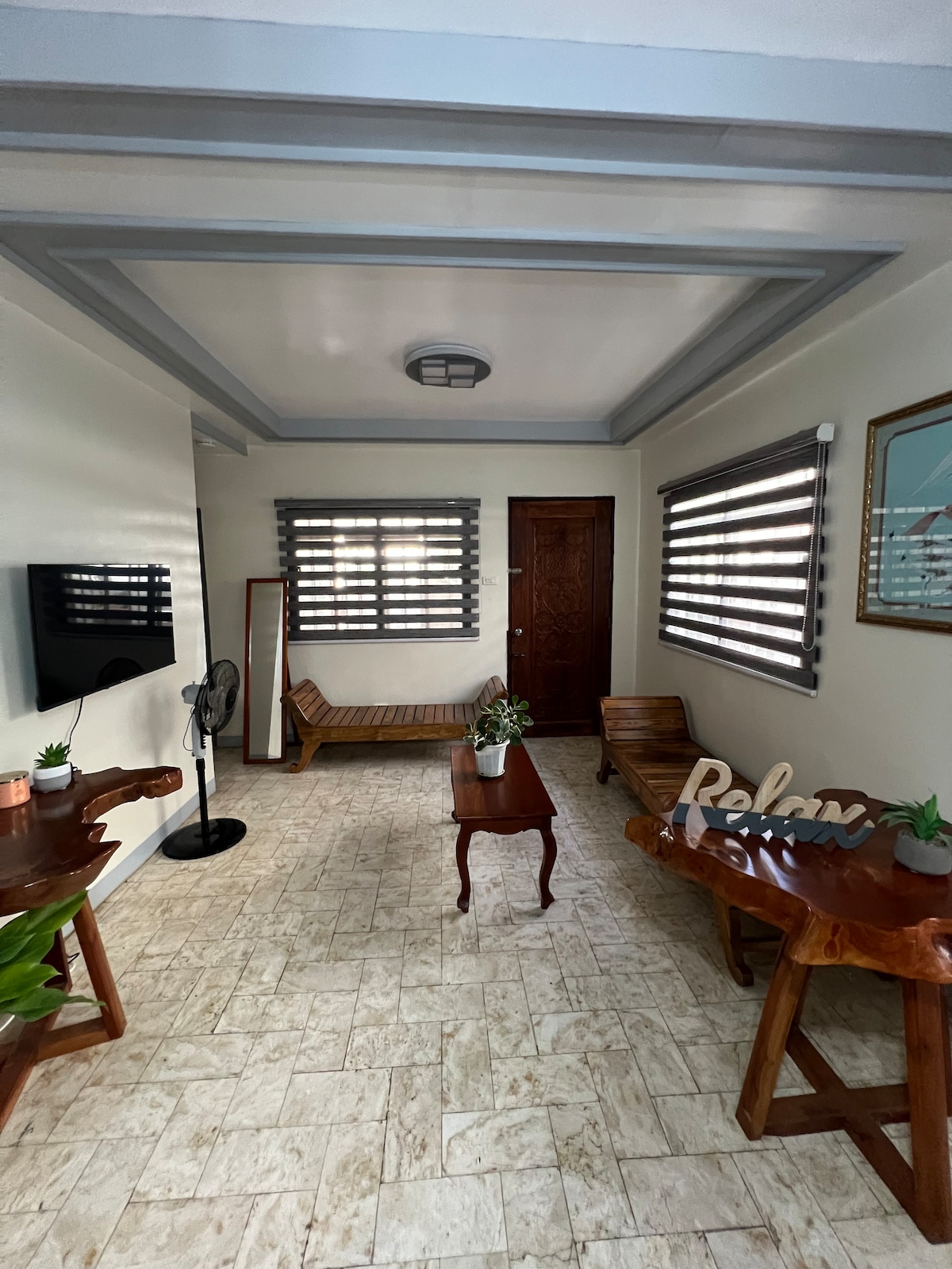 A welcoming living area features a combination of wooden furniture, including a bench and two side tables. Natural light filters through the horizontal blinds, creating a bright atmosphere. A wall-mounted television is positioned across from the seating. A fan and a decorative plant accent the space.