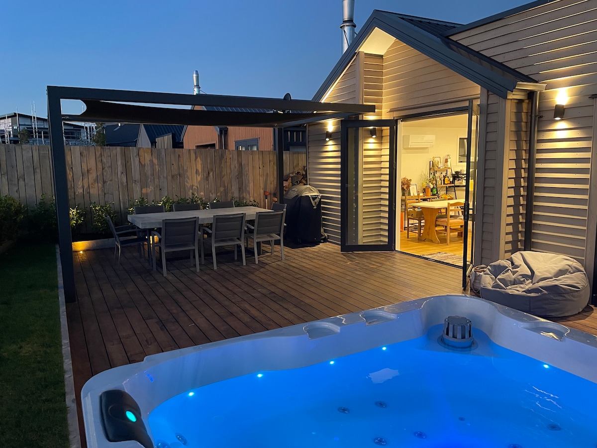An inviting outdoor space features a modern hot tub with illuminated water, bordered by a spacious wooden deck. A dining table is set nearby under a shaded area, while large glass doors open to the interior, creating a seamless flow between indoor and outdoor living.