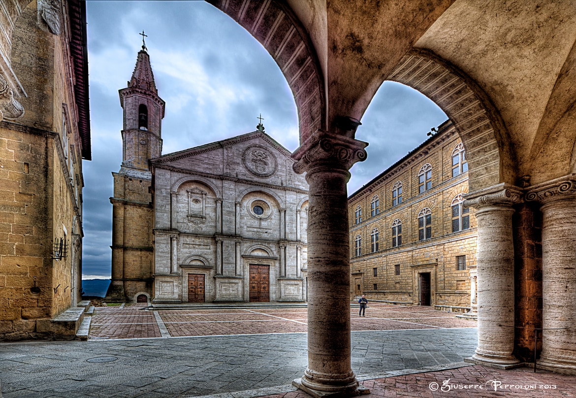 A tranquil plaza is featured, flanked by archways and historical stone buildings. The main church displays a simple yet elegant facade with a bell tower. Dim light filters through the clouds, creating a serene atmosphere in the square.